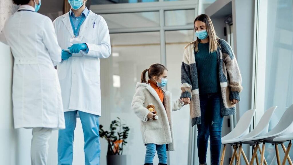 Masked mother holding her child’s hand in a clinic waiting area while two healthcare workers in white coats stand nearby.
