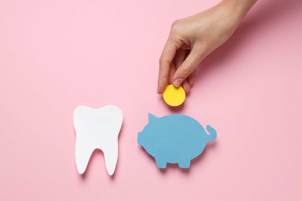 Hand placing a coin into a piggy bank beside a tooth icon, showing factors that affect Hollywood smile cost in Canada.