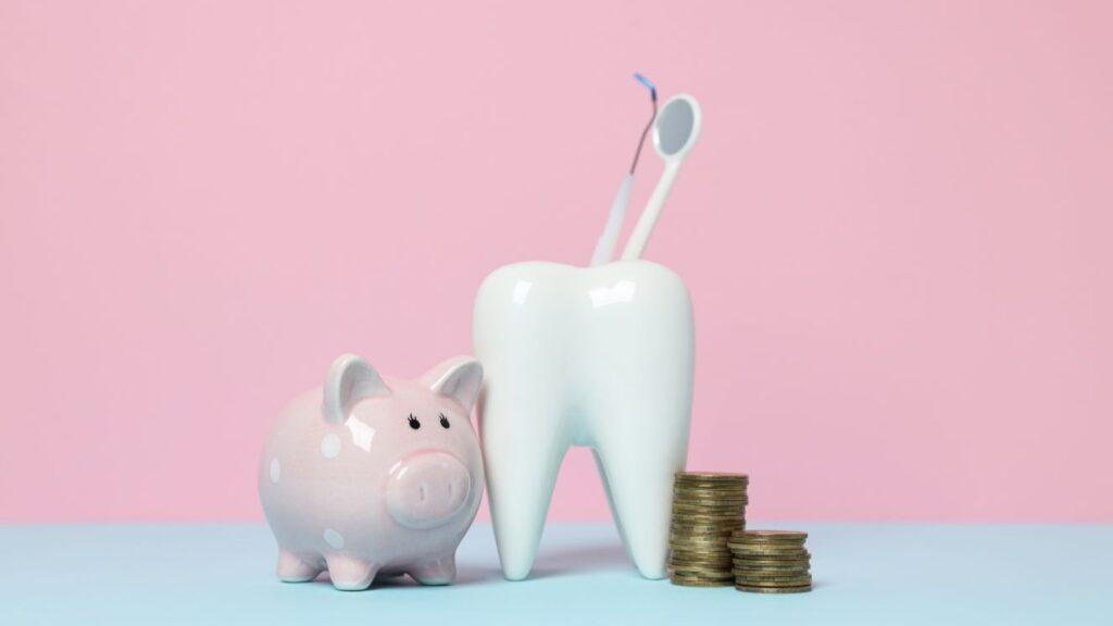 Pink piggy bank beside a large white tooth holding dental tools, with stacks of coins on a blue surface against a pink background.