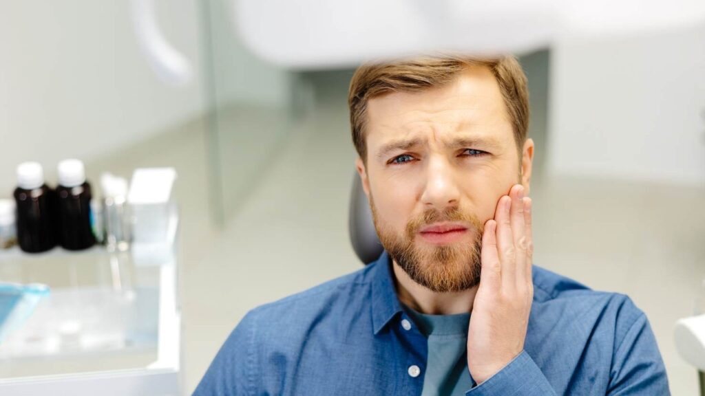 Man in a dental clinic holding his cheek in discomfort, representing possible sensitivity or pain after cosmetic dental treatment.