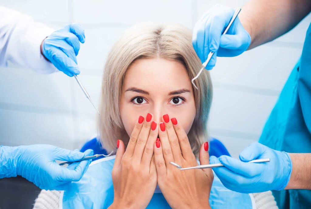 Anxious patient in a dental chair surrounded by gloved hands holding dental instruments, illustrating Hollywood smile risks and side effects.