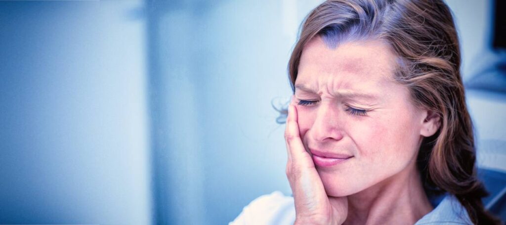 Woman with eyes closed pressing her cheek, showing facial/tooth pain, in a clinical setting.