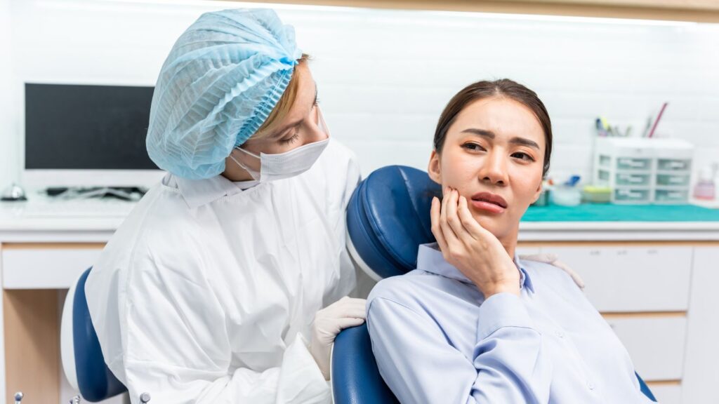 Patient holding her jaw with tooth pain while a dentist checks on her in a dental chair