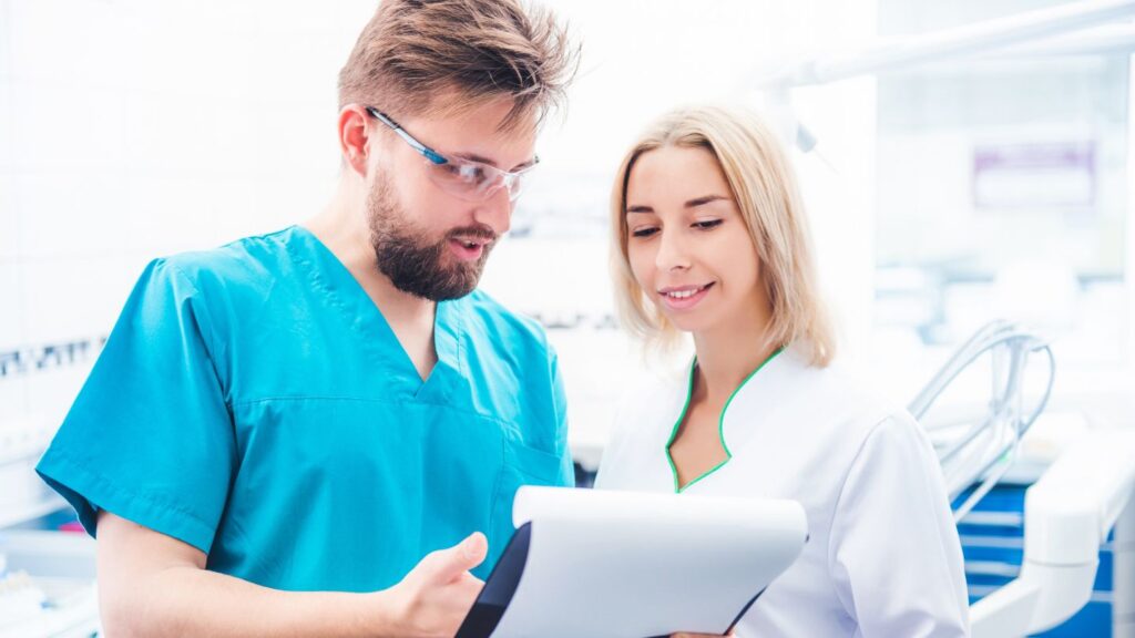 A dentist in blue scrubs and safety glasses reviews a treatment plan on a clipboard with a smiling woman in a dental clinic, representing how to compare Hollywood smile quotes and options.